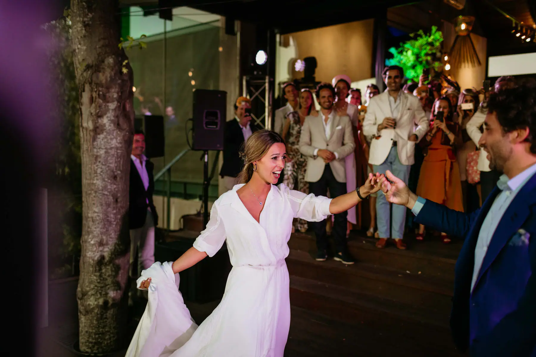 Barbara lifts her skirt and reaches for Mario's hand as guests with phones watch beside a speaker and light truss