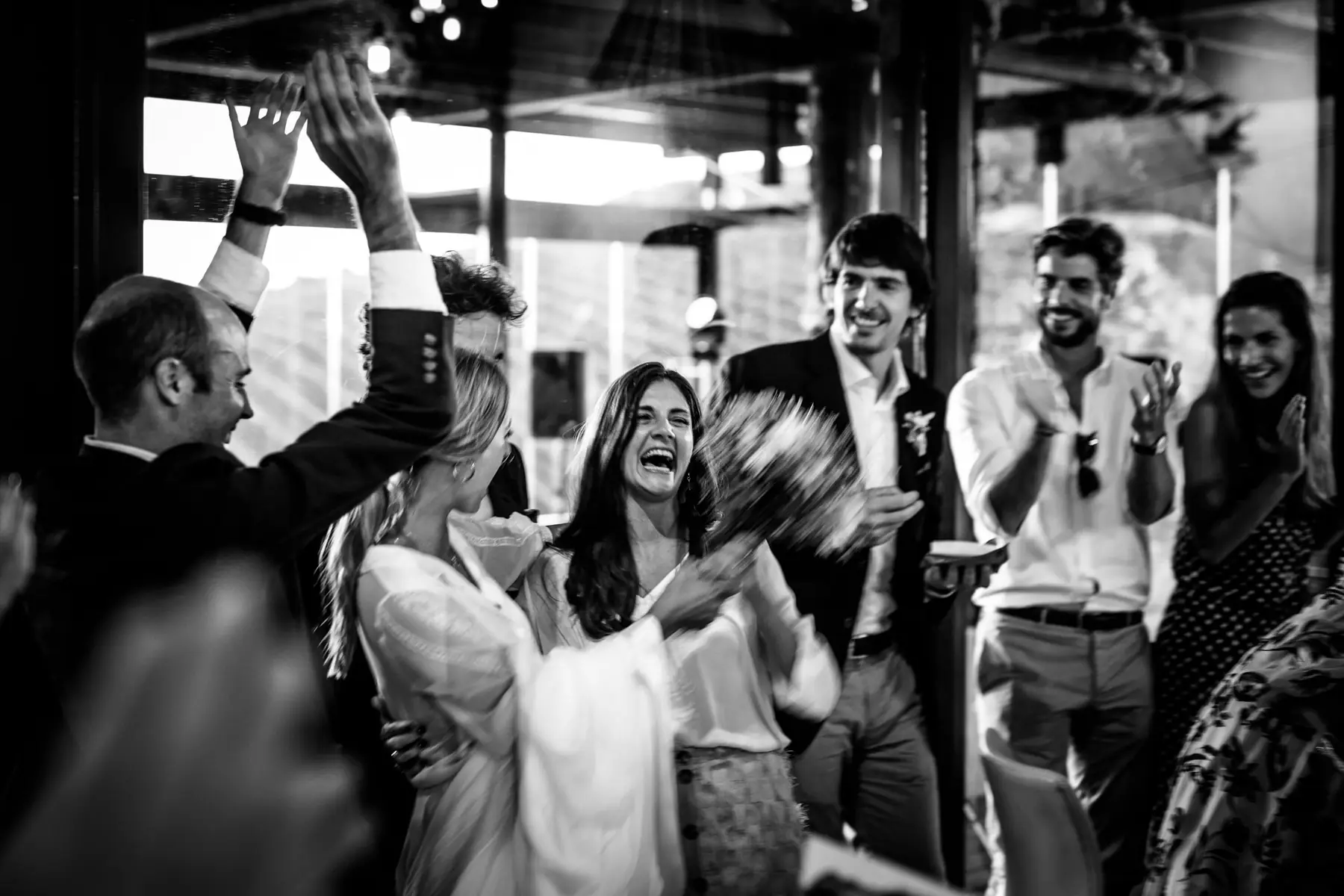 The bride hugs a laughing guest holding the bouquet as friends clap around them, in black and white