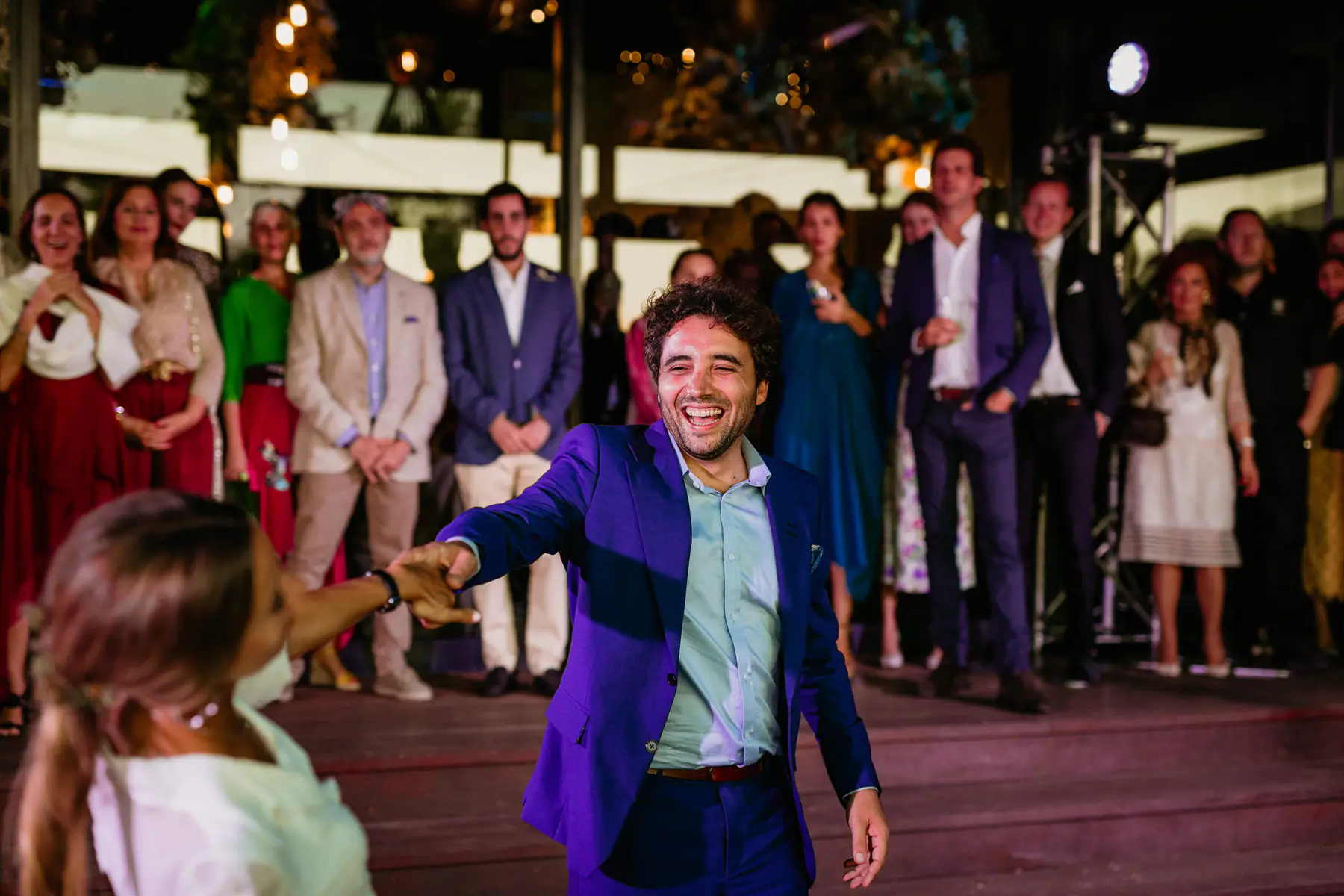 Mario, in a blue suit, laughs as he reaches out to lead Barbara into their first dance, guests lined up behind