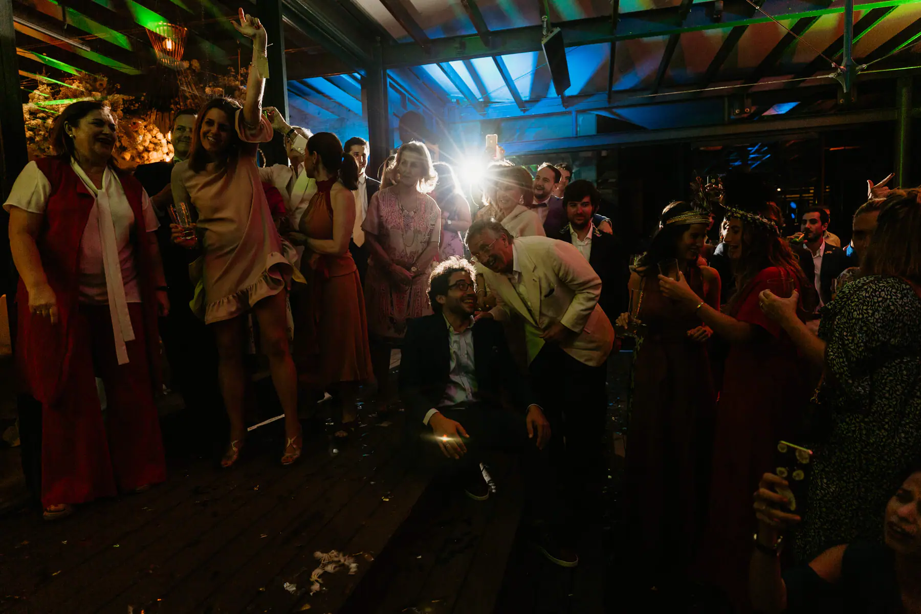 A man in glasses sits laughing on the deck steps as an older guest in a cream jacket leans over him and guests cheer