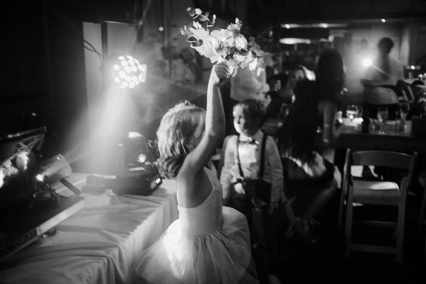 A flower girl lifts the bouquet beside a moving head light on the DJ table as a young boy watches, in black and white