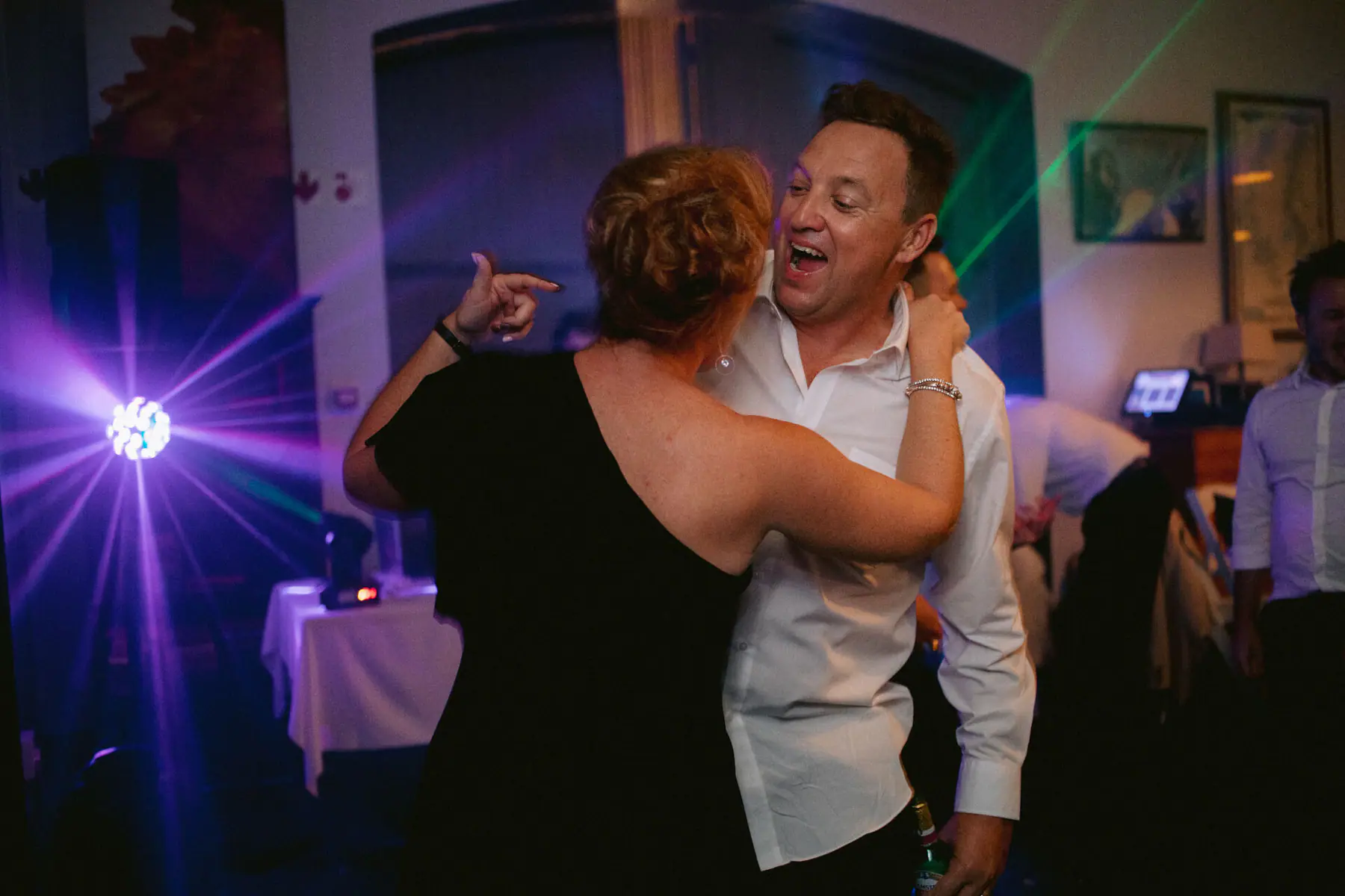 A guest in a white shirt sings as he dances with a woman in a black one-shoulder dress beside the DJ table's purple light