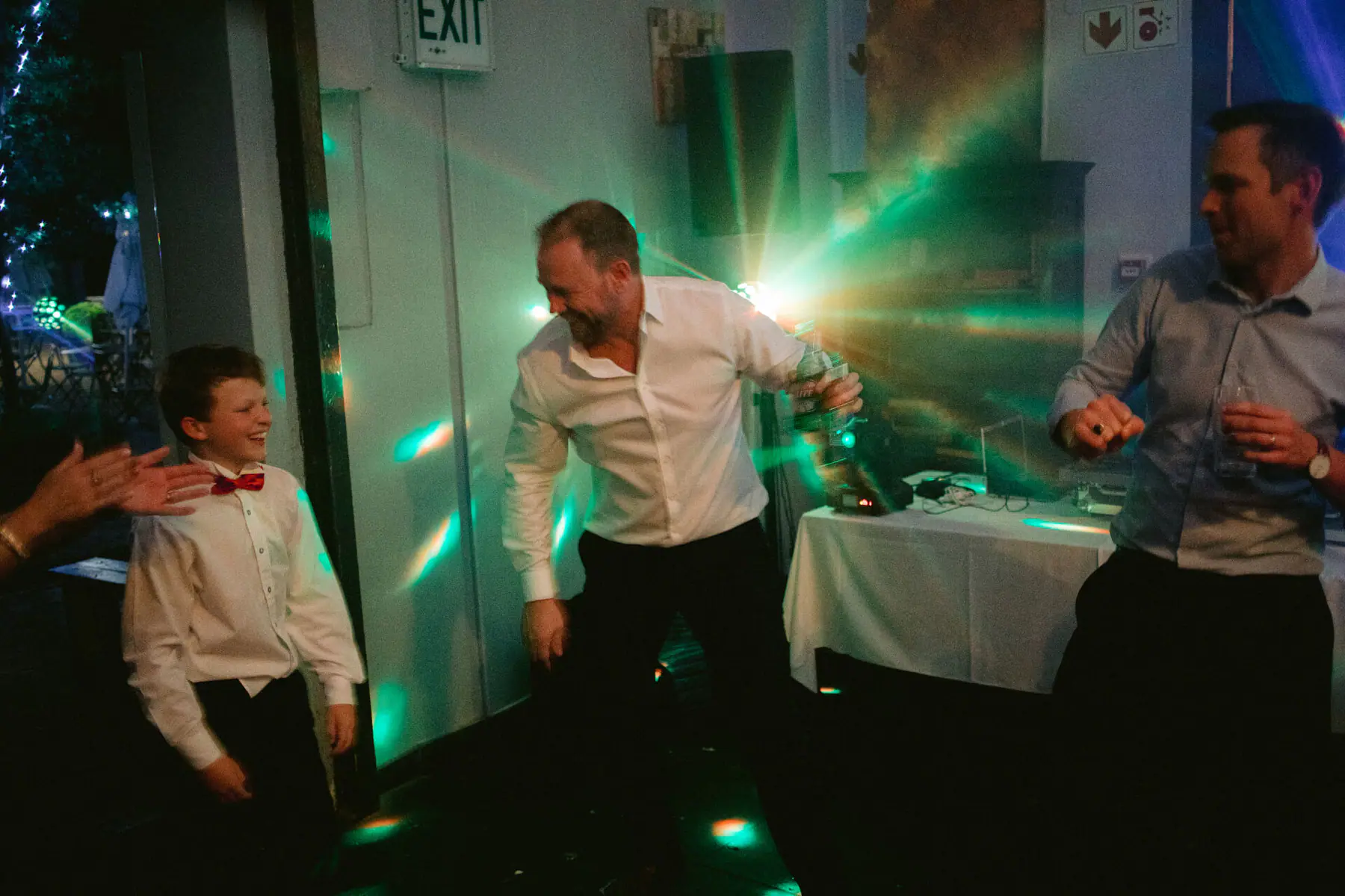 A young guest in a red bow tie laughs as a man in a white shirt dances by the DJ table under green disco beams