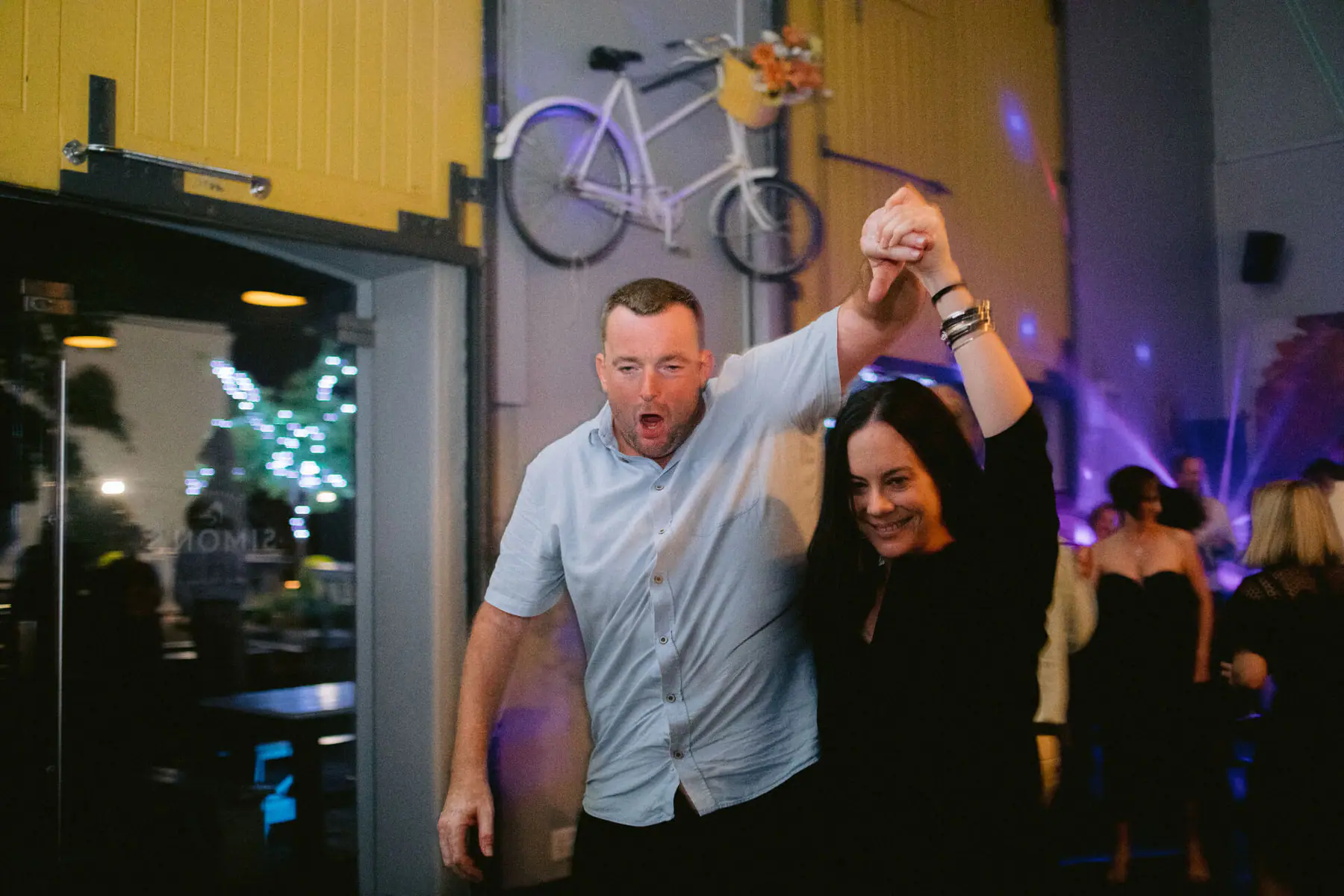 A guest in a pale blue shirt twirls a partner in black below a white bicycle mounted on a yellow barn door in Constantia