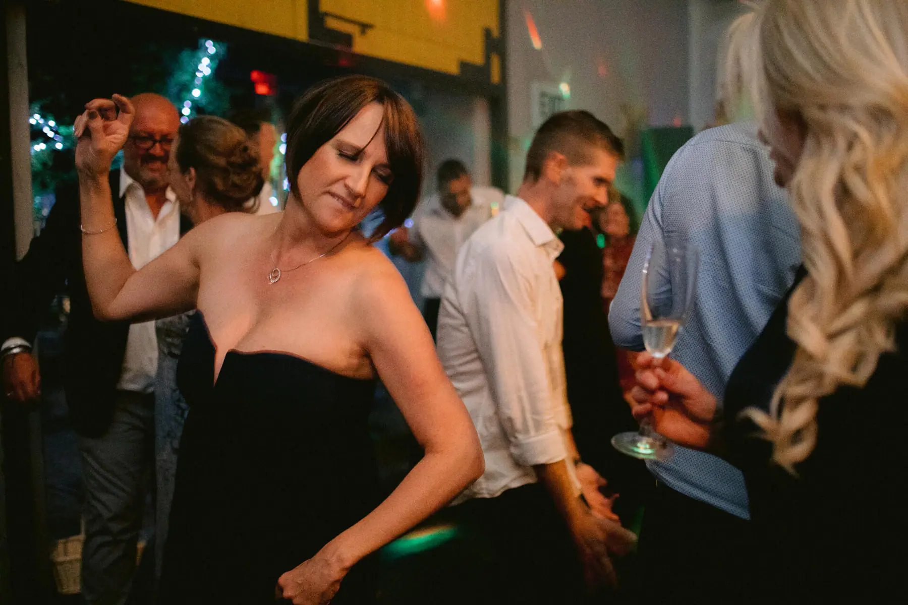 A guest in a black strapless dress dances with her eyes closed as a friend holds a champagne flute in the foreground