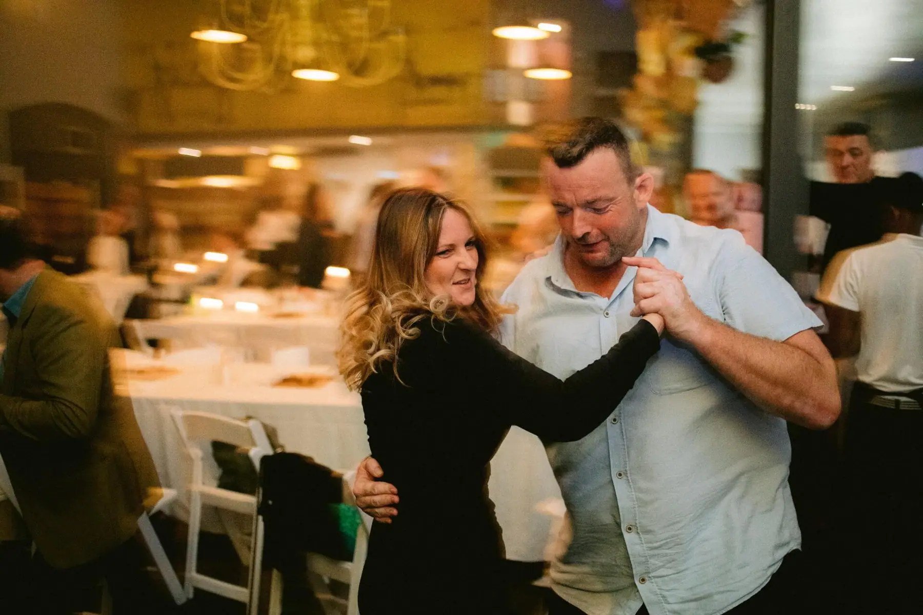 Two guests hold hands in a partner dance between white dining tables under warm pendant lights at Simon's