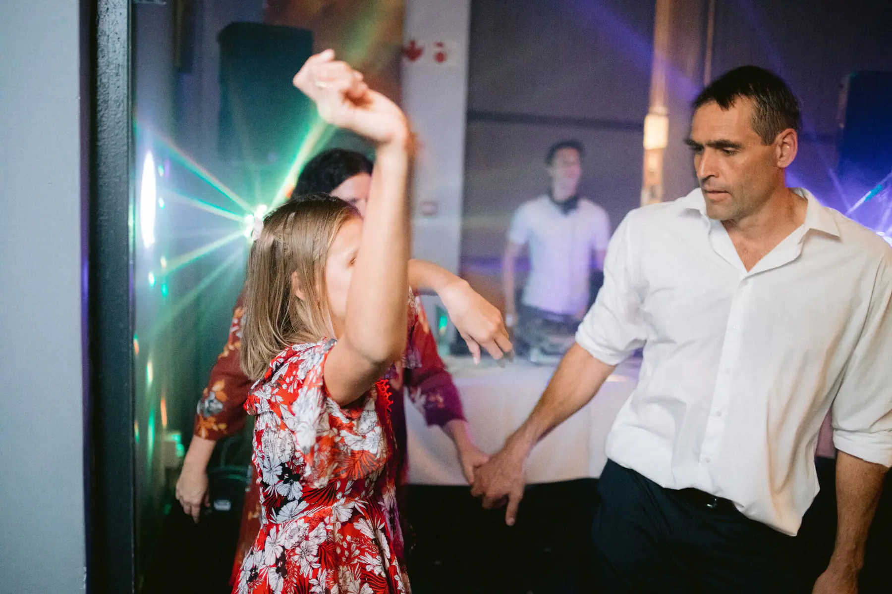 A young guest in a red floral dress dances beside a man in a white shirt while the DJ plays behind them in green light