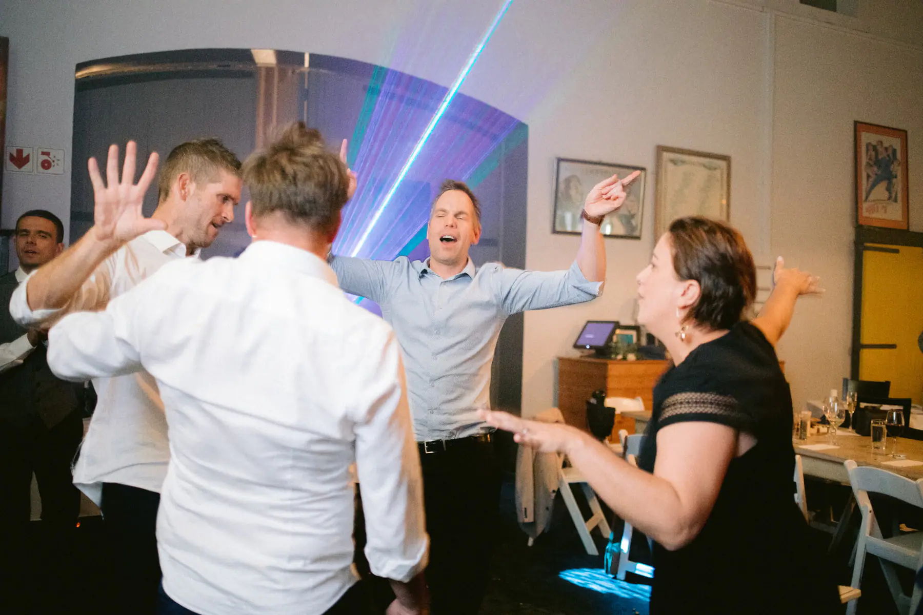 Guests dance with hands up as a man in a light blue shirt sings, arms wide, in front of blue laser beams