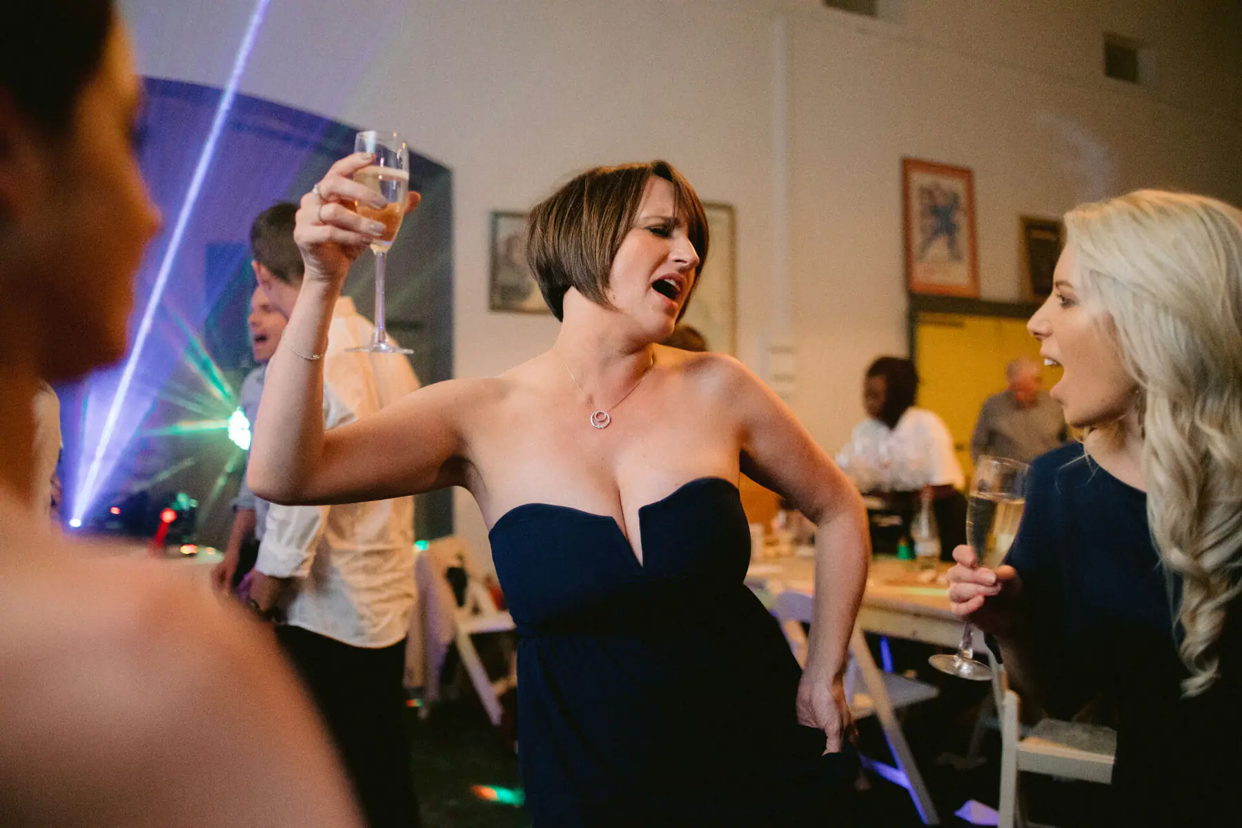 A guest in a navy strapless dress sings with her champagne flute raised as a friend laughs and a laser beam crosses the room