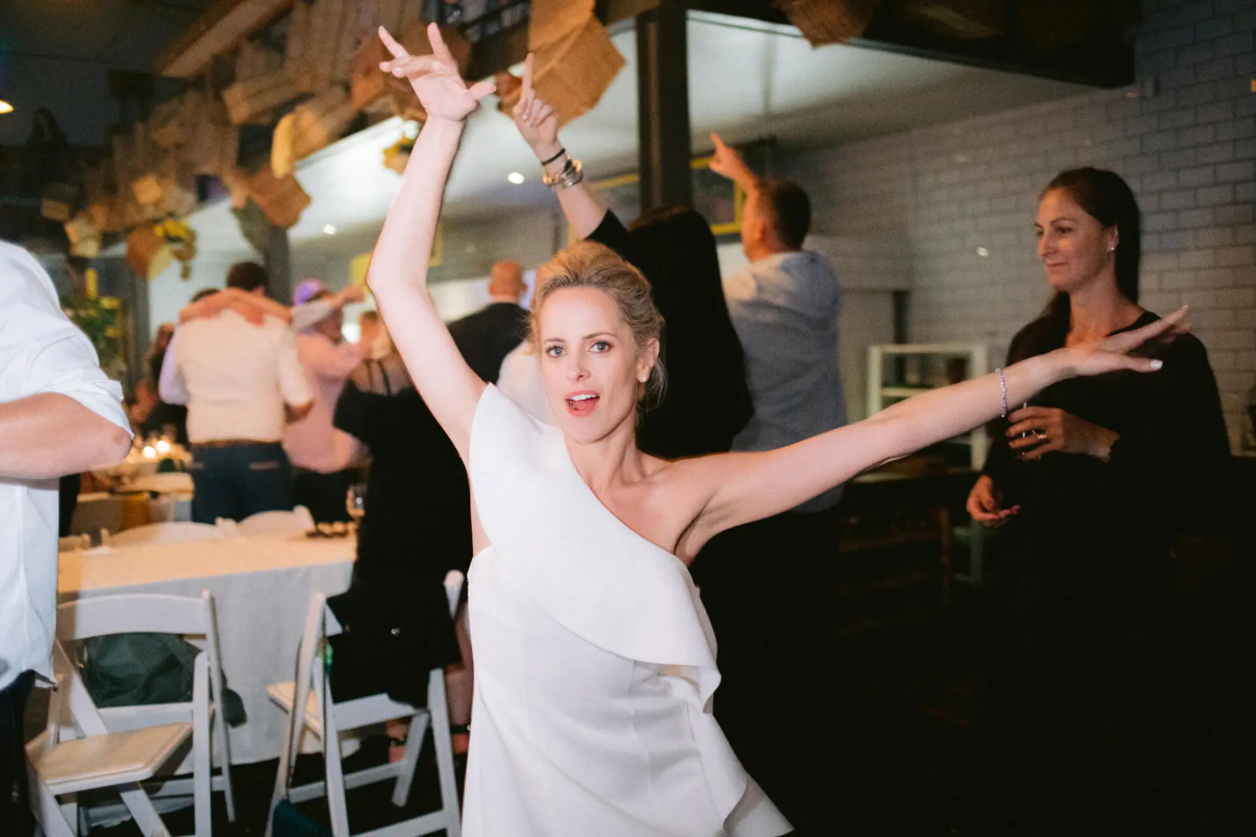 Kirsten throws both arms up as she dances among guests beside dining tables, wooden crates hanging from the ceiling
