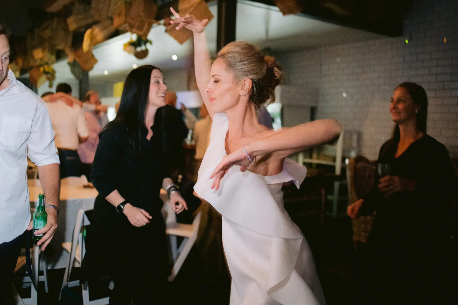 Kirsten dances with one hand raised high as a guest in black laughs and a man holds a beer beside the dining tables