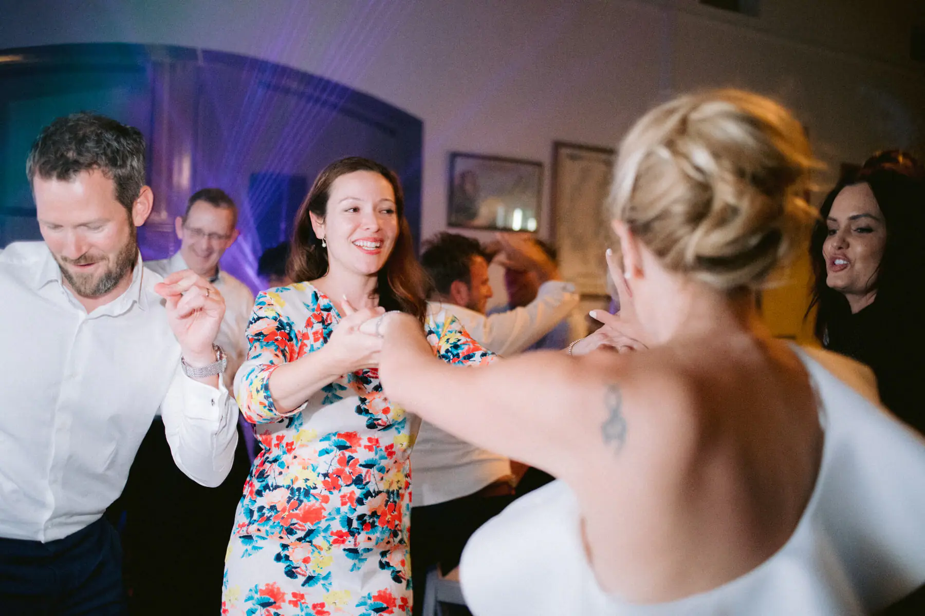 Kirsten, seen from behind, holds hands with a guest in a floral dress as friends dance around them in purple light