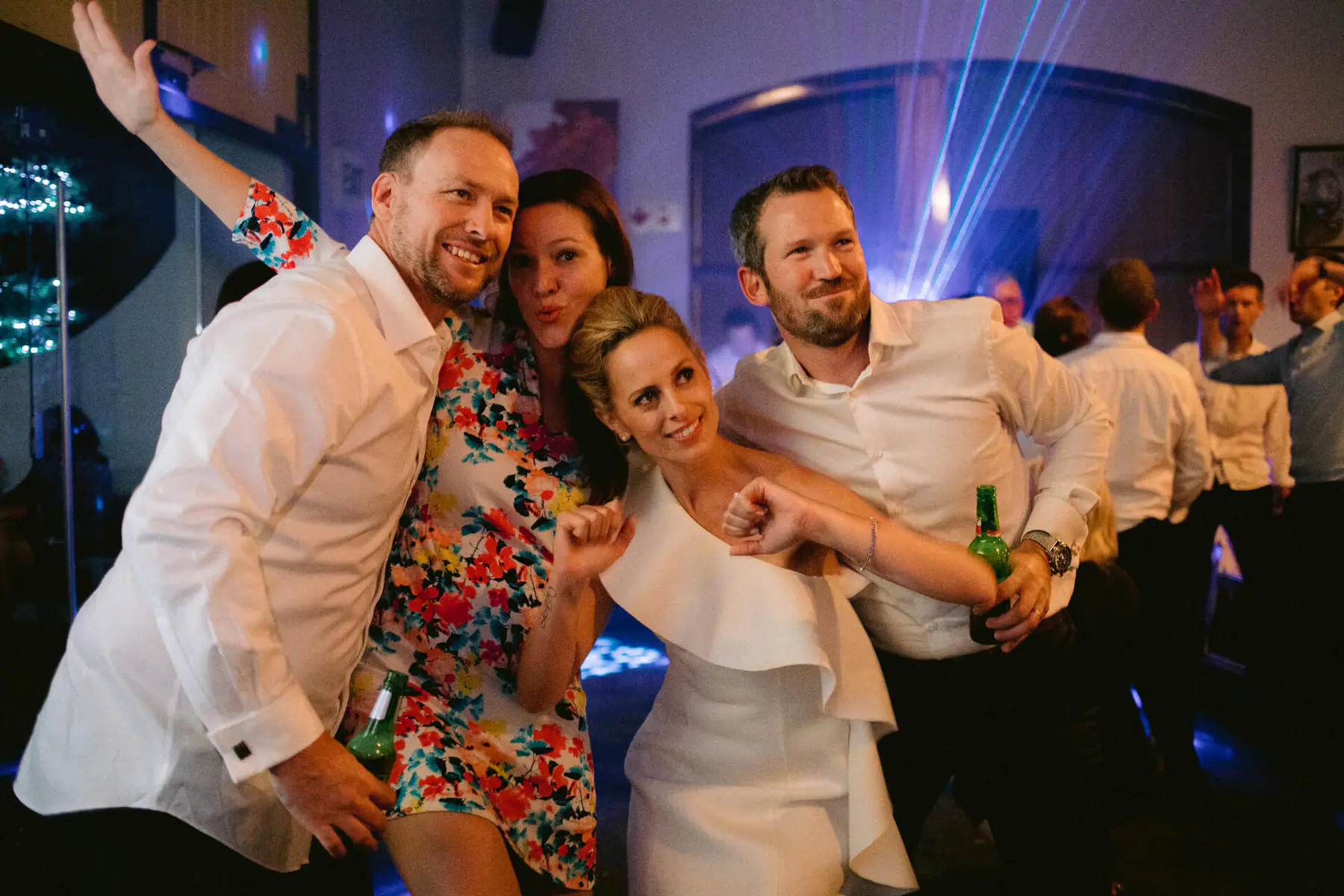 Kirsten and Greg pose with two friends, fists raised and beers in hand, as blue laser beams fan across the wall