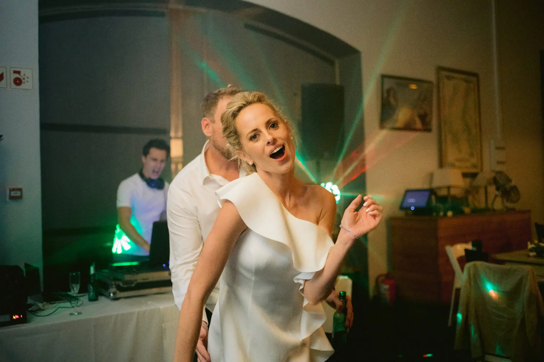 Kirsten sings towards the camera with Greg behind her as the DJ in headphones plays at the booth under green lasers