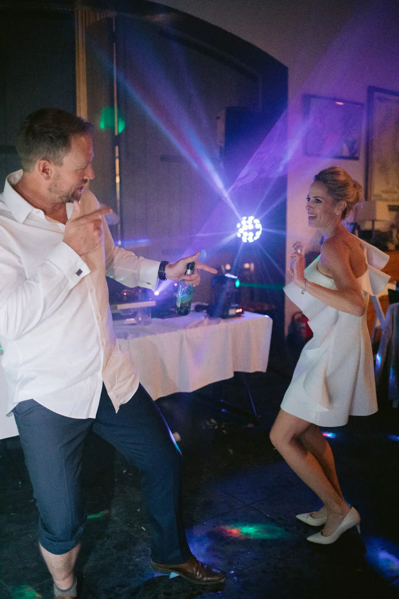 Kirsten in a white ruffled dress and heels dances opposite Greg in front of The DJ Company's purple-lit DJ table