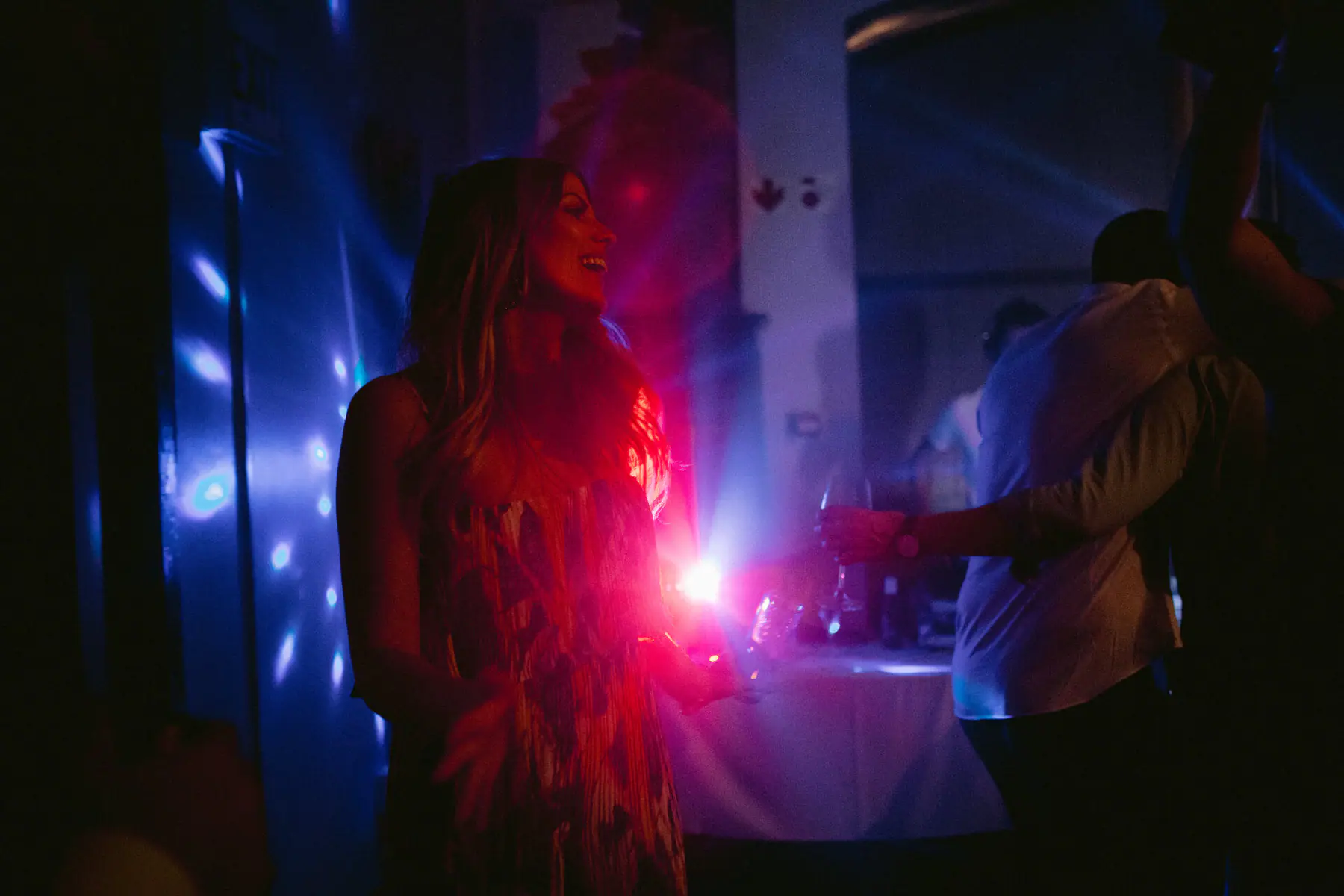 A guest in a patterned dress laughs in the dark, lit pink and blue by party lights throwing dots across the wall