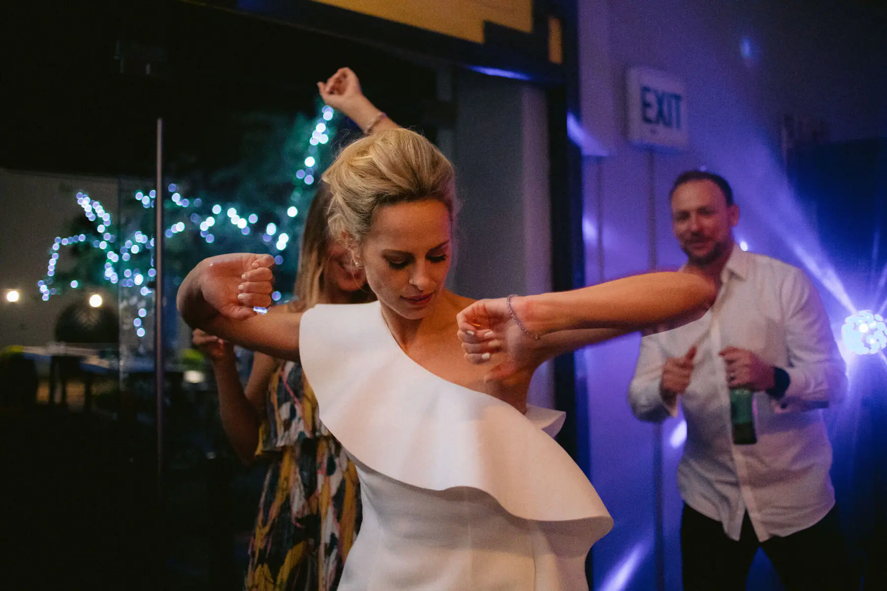 Kirsten dances with her elbows raised as Greg watches with a beer, a fairy-lit tree glowing through the glass doors