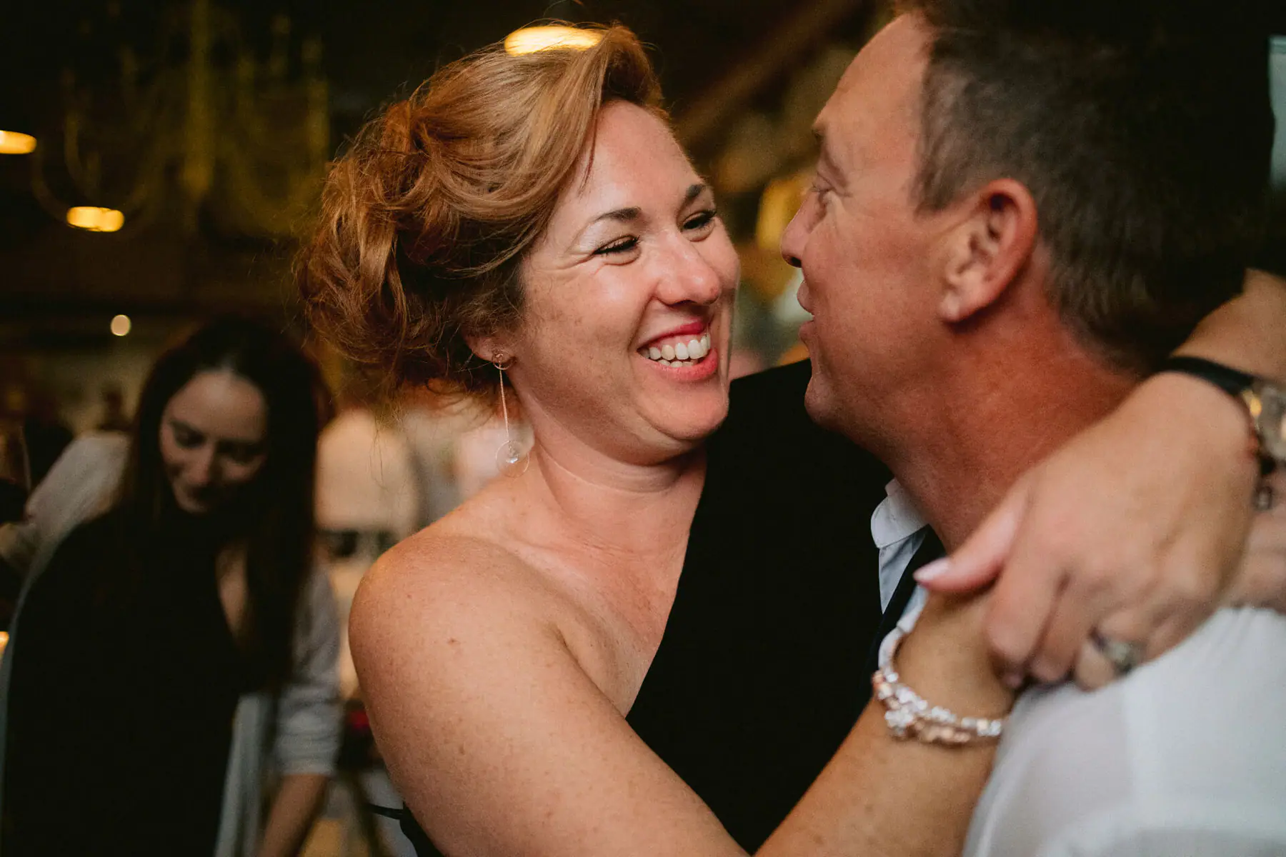 A guest with auburn hair laughs as she hugs a man close on the dance floor, warm lights behind at Groot Constantia