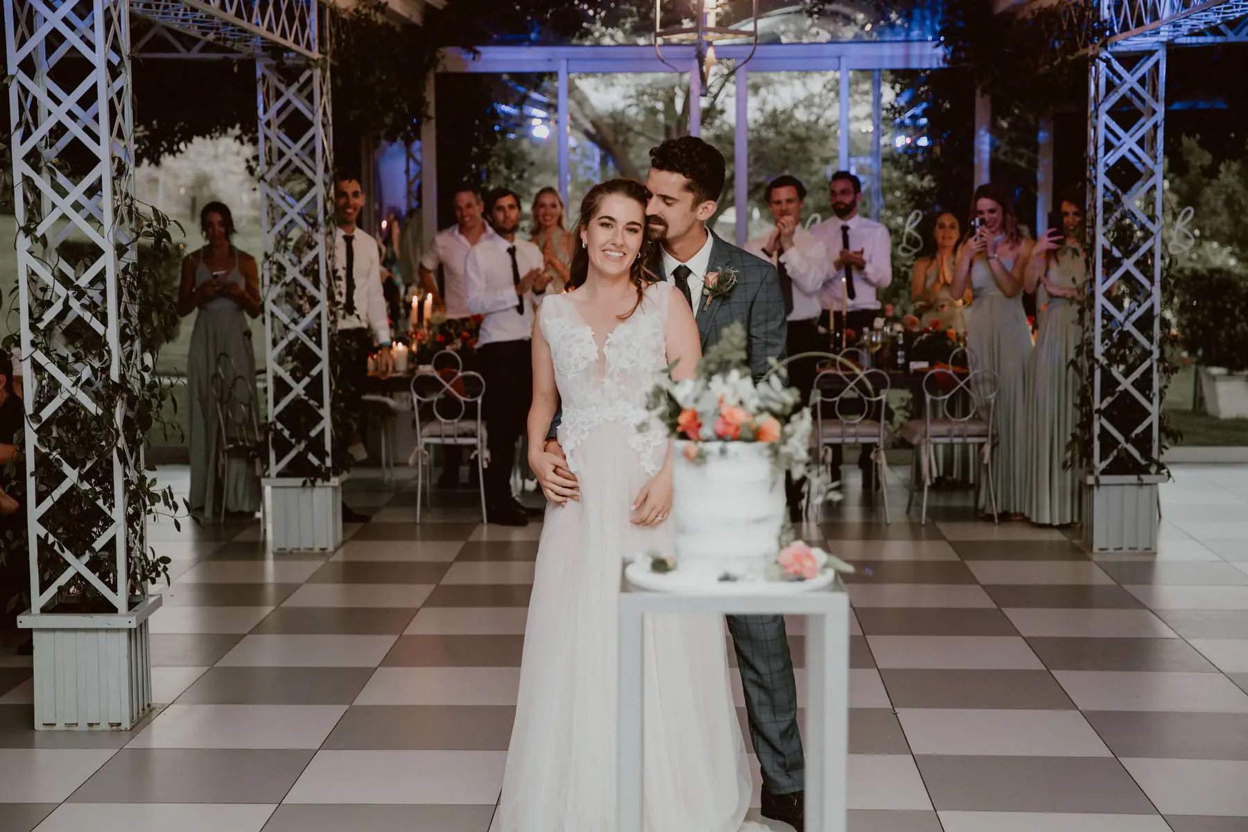 Madison and Karel behind a semi-naked wedding cake topped with peach roses as groomsmen and bridesmaids clap