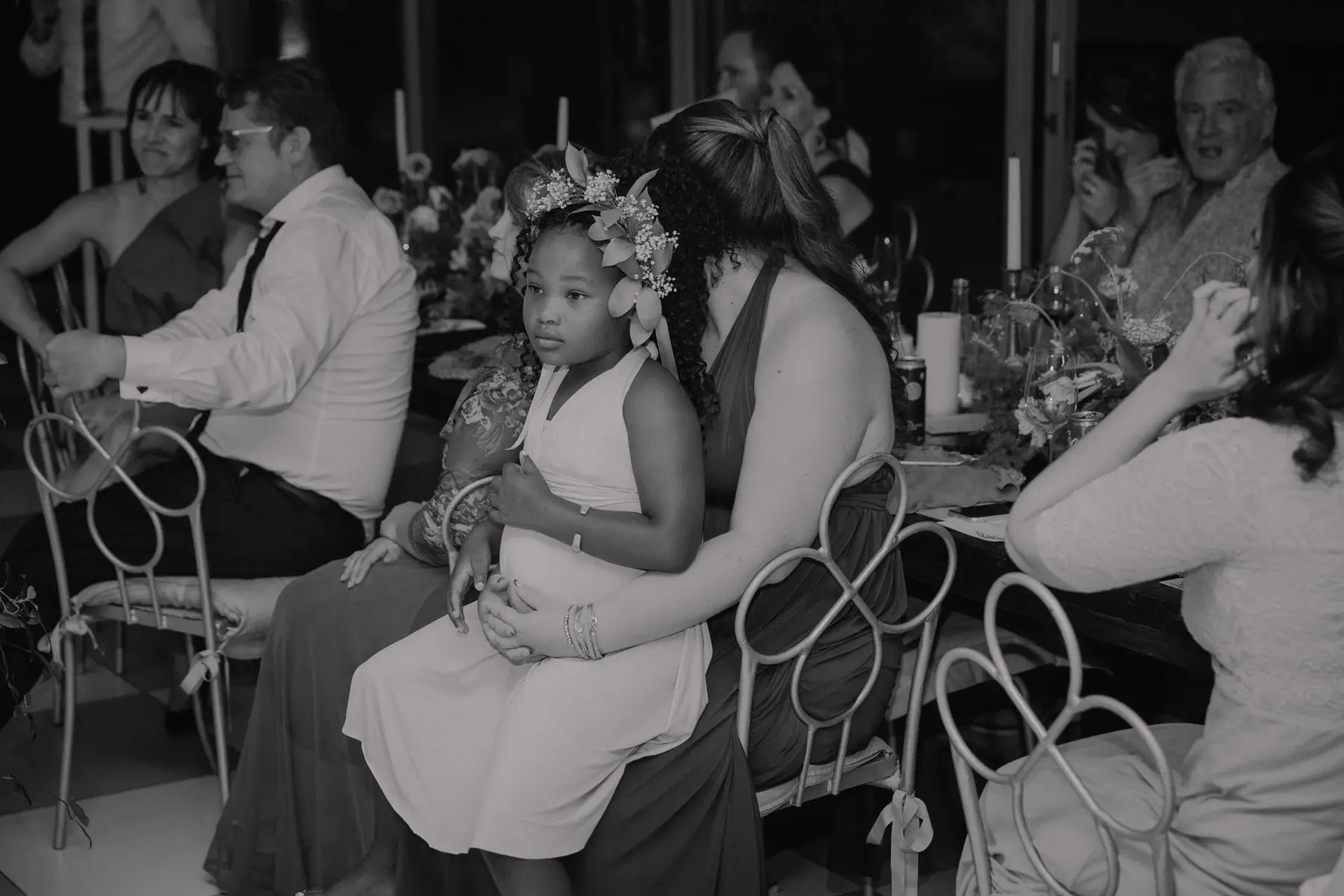 In black and white, a flower girl in a floral crown sits on a bridesmaid's lap as guests at the candlelit table look on