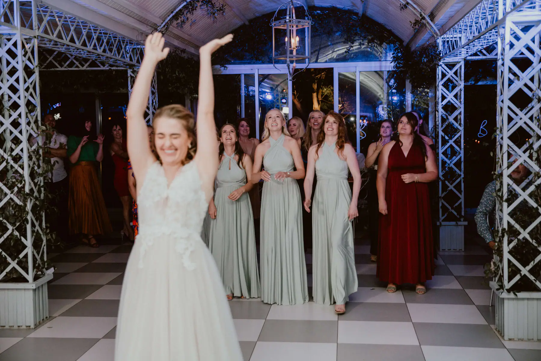 Madison with both arms in the air after tossing her bouquet to bridesmaids in sage green gowns under a hanging lantern