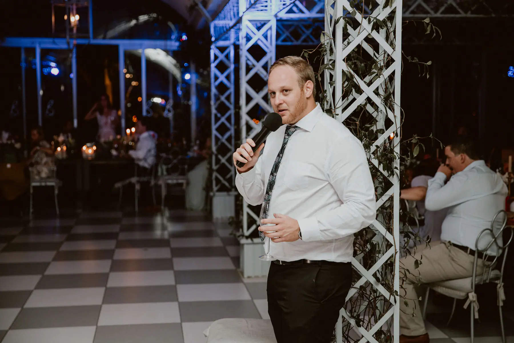 Guest in a patterned tie giving a speech on a handheld microphone beside an ivy-wrapped lattice pillar at Belair Pavilion