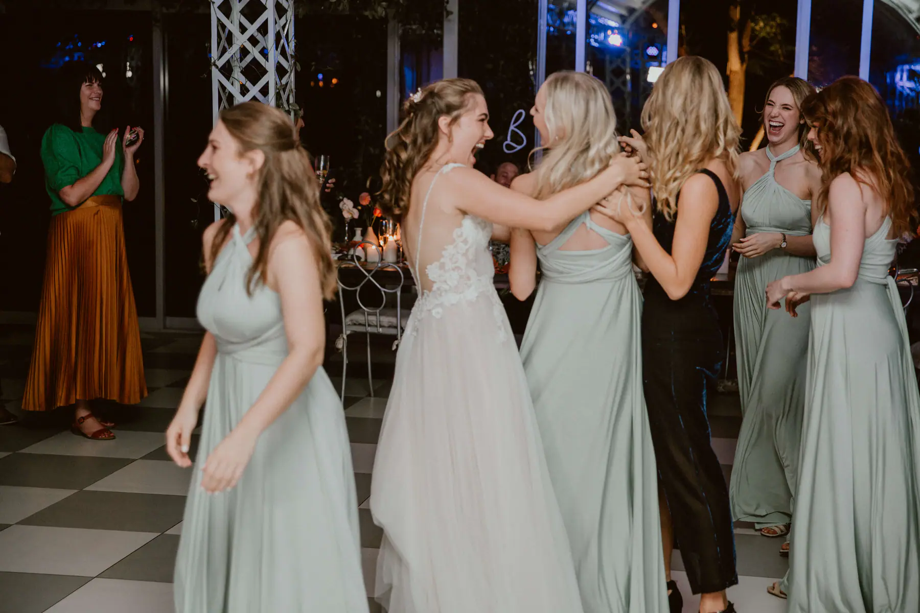 Madison laughing as she hugs bridesmaids in sage green halter gowns while a guest in a pleated orange skirt claps