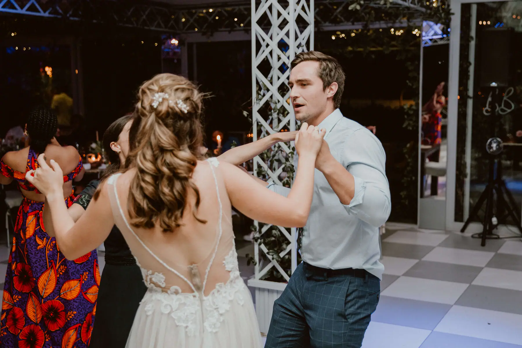 Madison seen from behind in a sheer lace-backed gown, dancing with a guest in a pale blue shirt who sings along