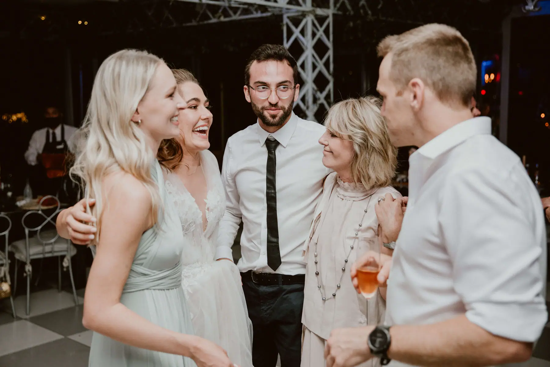Madison laughing in a huddle with a bridesmaid, a bearded guest in glasses and an older woman holding a glass of rosé