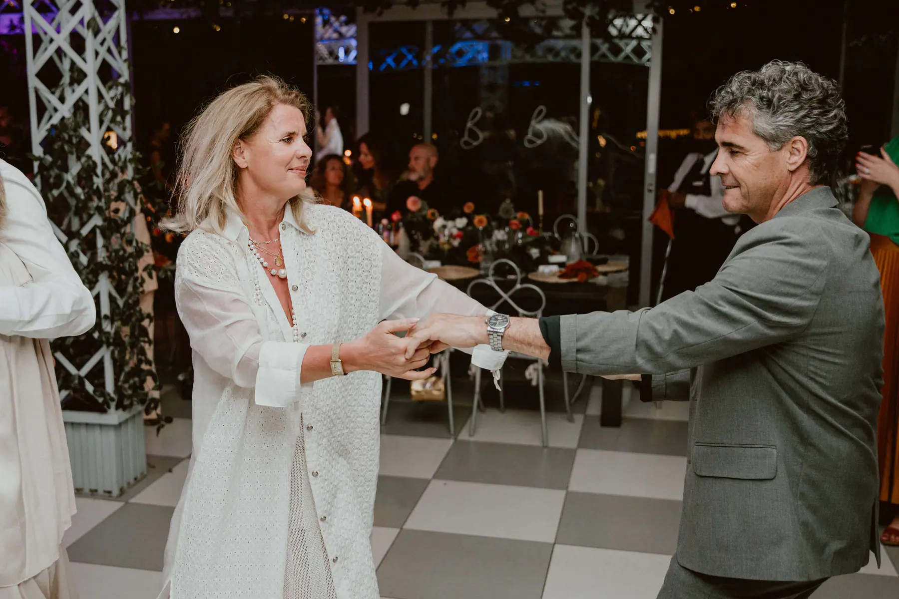 Woman in white lace and pearls dancing hand in hand with a grey-haired man in a grey suit beside candlelit tables