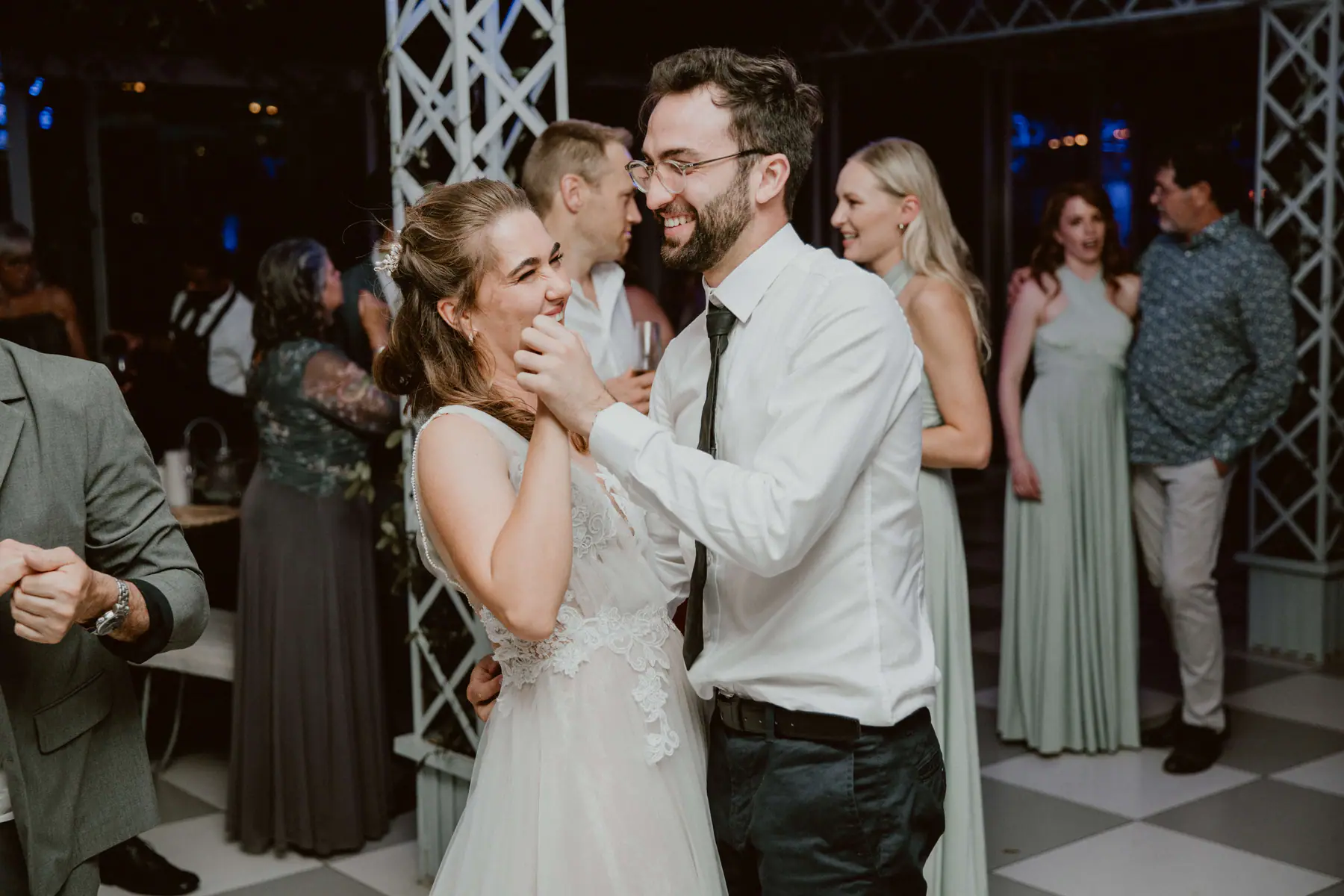 Madison laughing as a bearded guest in glasses and a black tie holds her hands mid-dance, bridesmaids in sage behind