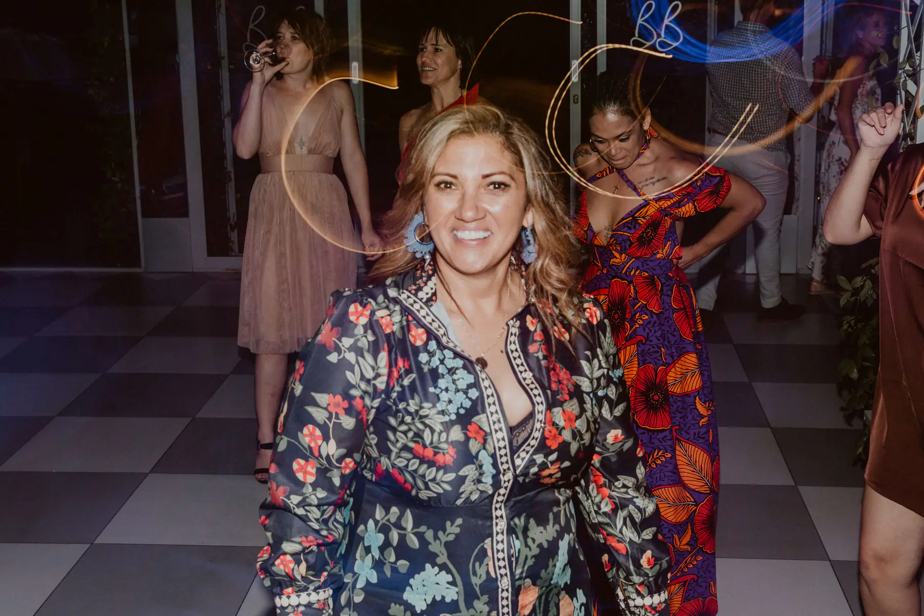 Smiling guest in a navy floral dress and blue tassel earrings, dance floor light trails swirling behind her