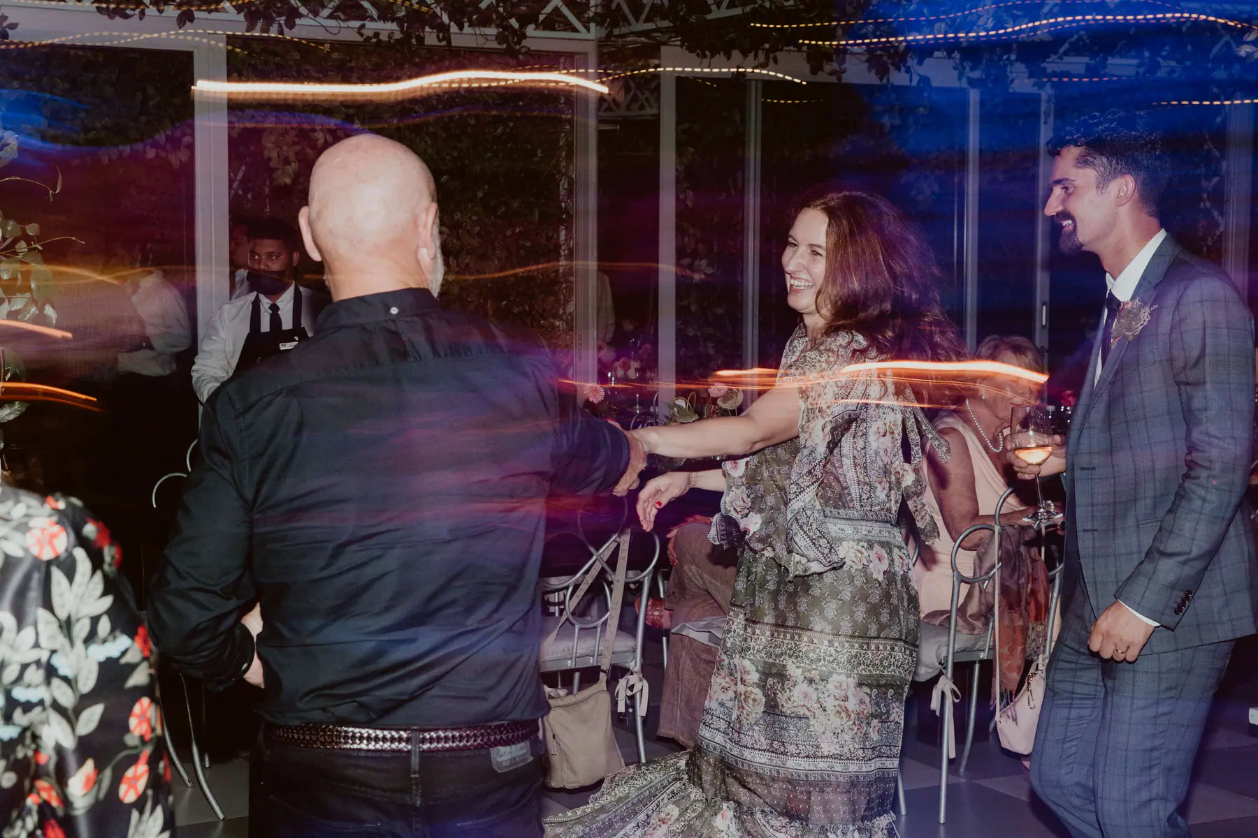 Karel watching with a glass of wine as a bald guest in a black shirt dances with a woman in an olive print dress