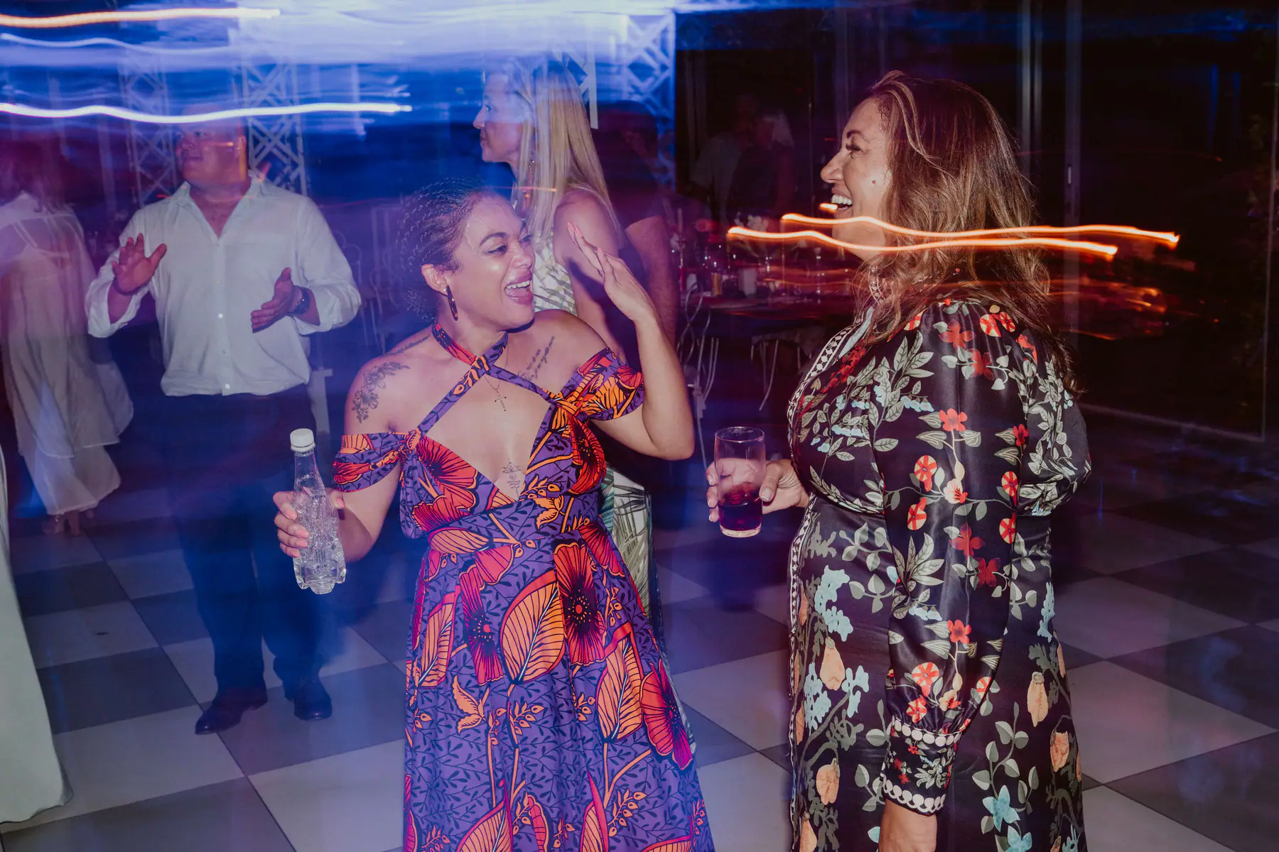 Two guests laughing on the dance floor, one in a purple and orange wax print dress, amid streaks of orange light
