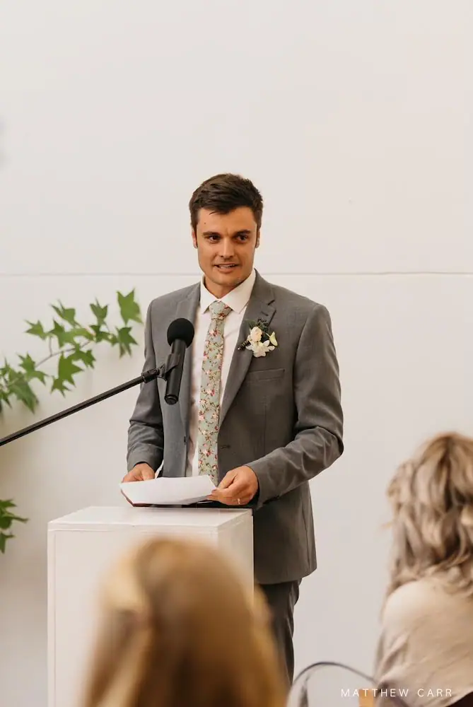 Man in a grey suit, floral tie and buttonhole giving a wedding speech at a white lectern with a microphone