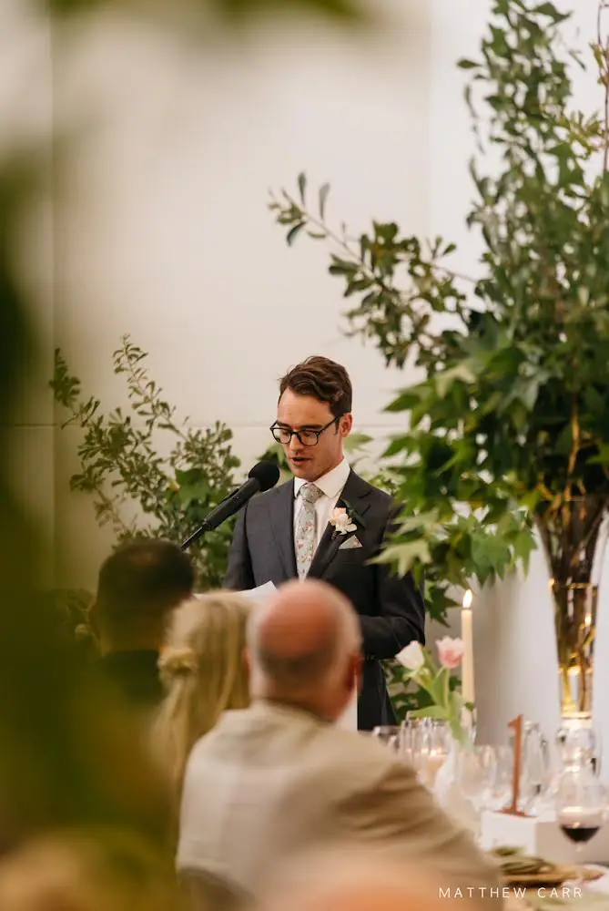 Man in glasses and a floral tie reading a speech into a microphone beside greenery, candles and pink tulips