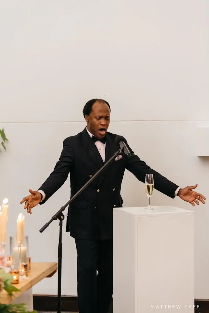 Singer in a double-breasted dinner jacket and bow tie performing with arms outstretched at a microphone beside candles