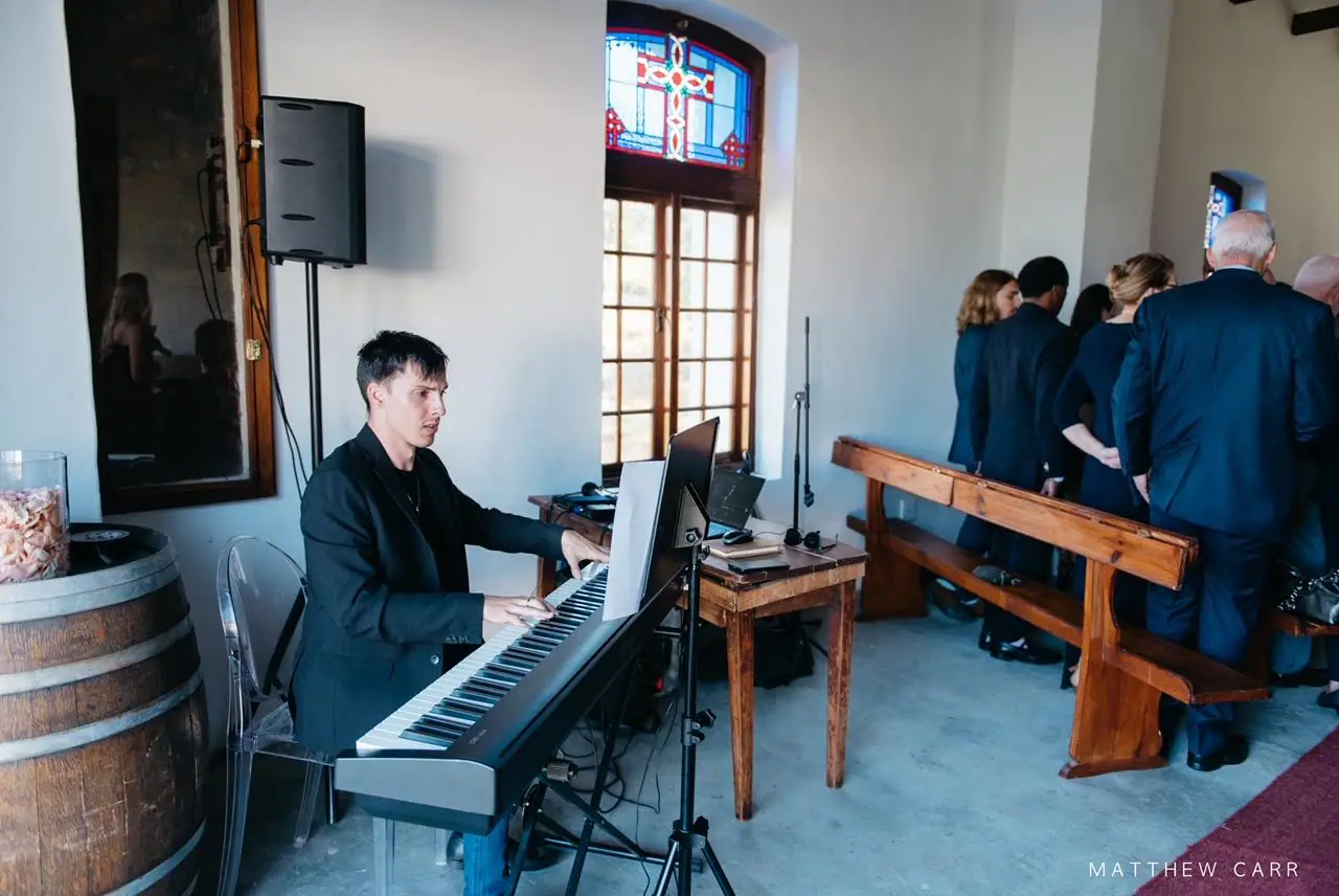 Pianist at a digital piano in a whitewashed chapel with a stained glass window, wooden pews and guests standing