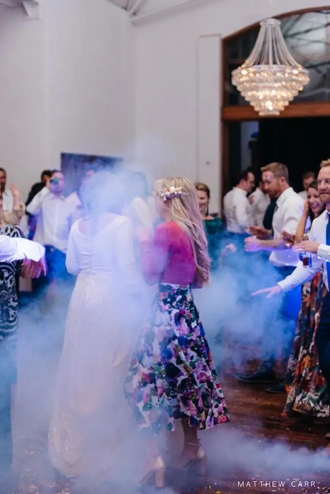 Camilla in her sequinned gown dancing with a guest in a floral skirt through thick haze under a crystal chandelier