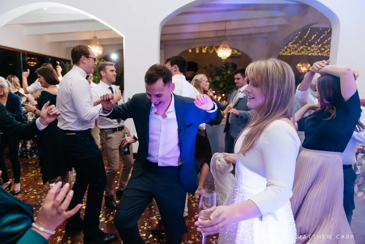 Camilla laughing with a glass of wine as a guest in a blue blazer dances beside her on the confetti-covered floor