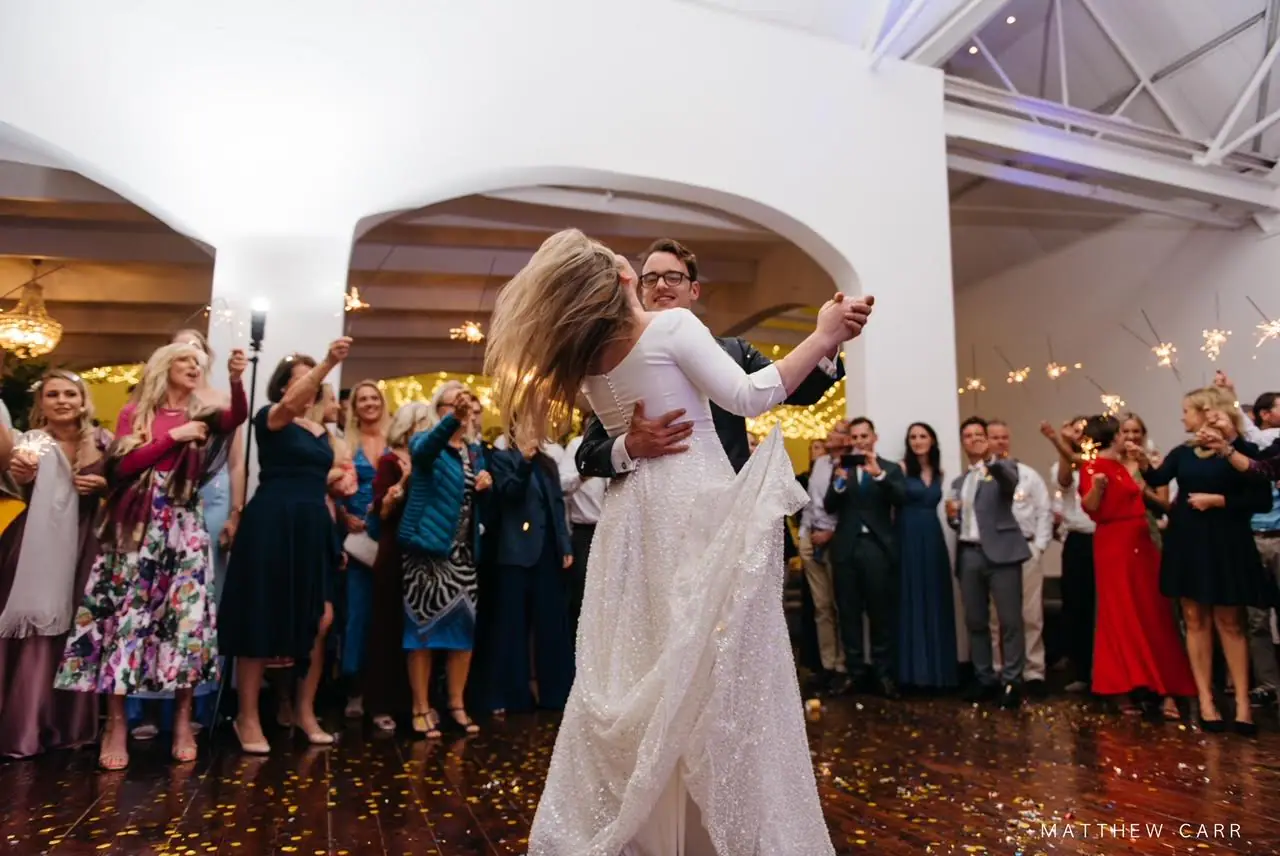 Nick holding Camilla close in their first dance, her hair swinging, as guests with sparklers ring the wooden floor