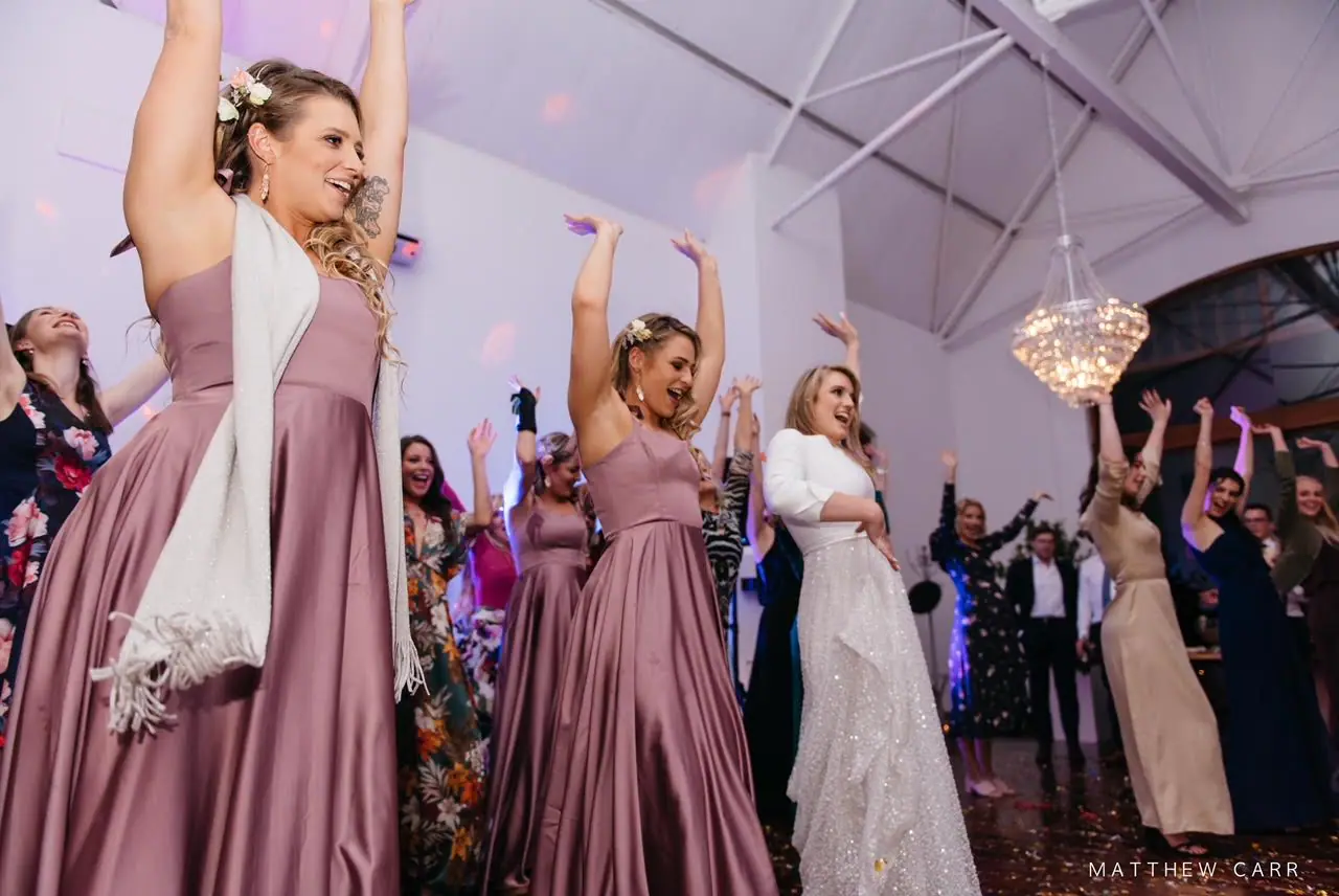 Camilla and her bridesmaids in dusty pink satin gowns dancing with their arms up under a crystal chandelier