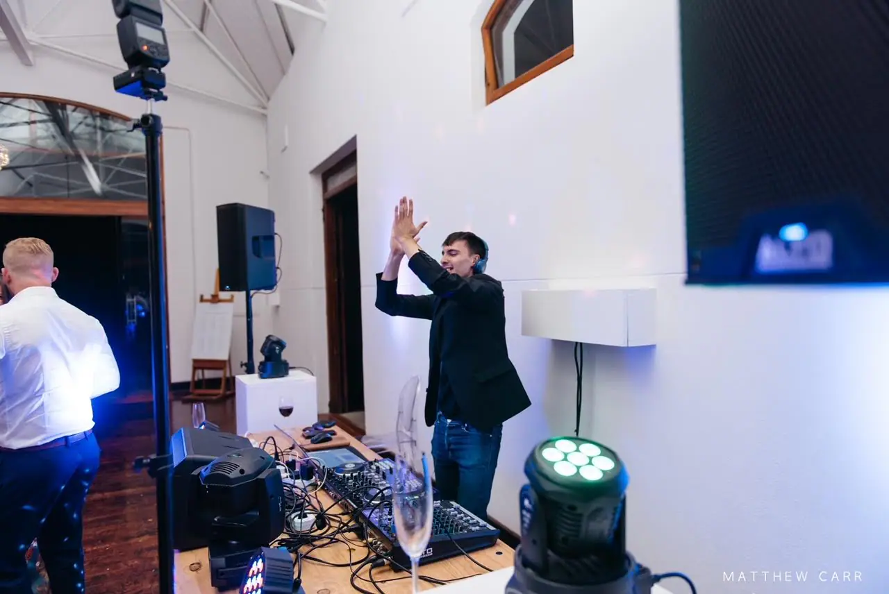 DJ clapping his hands above his head at the decks, with a green moving head light and a speaker on a stand nearby