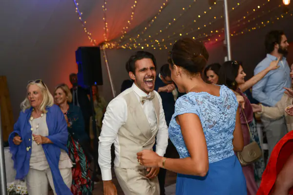 Groom in a cream waistcoat and bow tie laughing as he dances with guests under marquee fairy lights