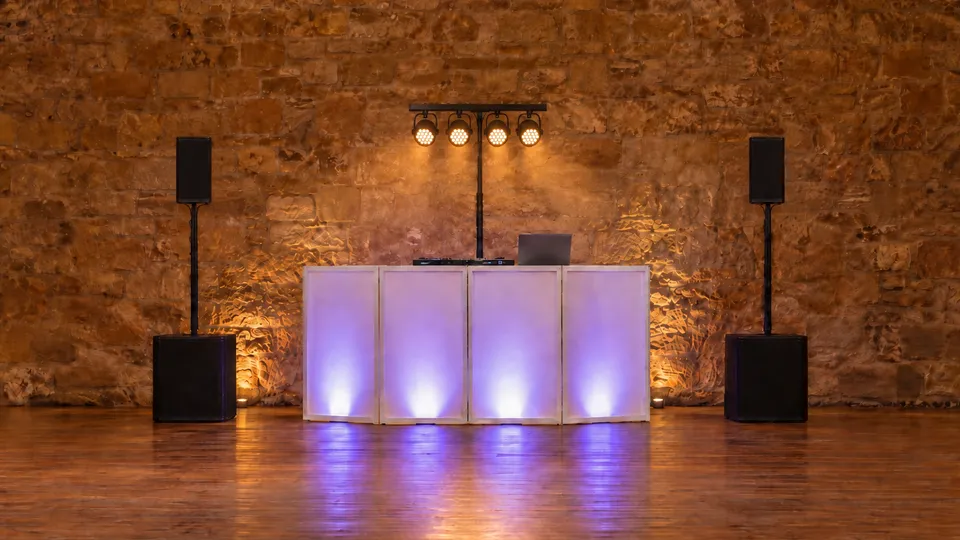 Dance floor setup in a sandstone barn: a white DJ facade lit violet from behind, a short T-bar with four PAR cans, and a speaker on a subwoofer either side