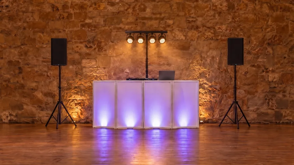 Dance floor setup in a sandstone barn: a white DJ facade lit violet from behind, a short T-bar with four PAR cans, and a speaker on a tripod stand either side
