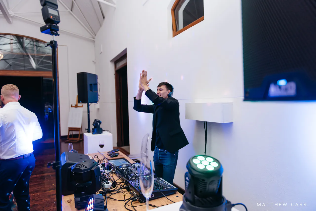 Liam at the DJ booth with his hands in the air beside a speaker and a moving head light
