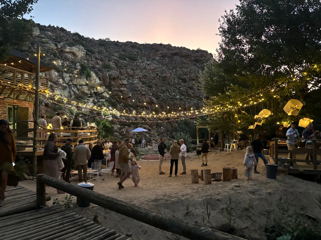 Wedding guests on an outdoor dance floor under string lights at dusk, below a rocky hillside